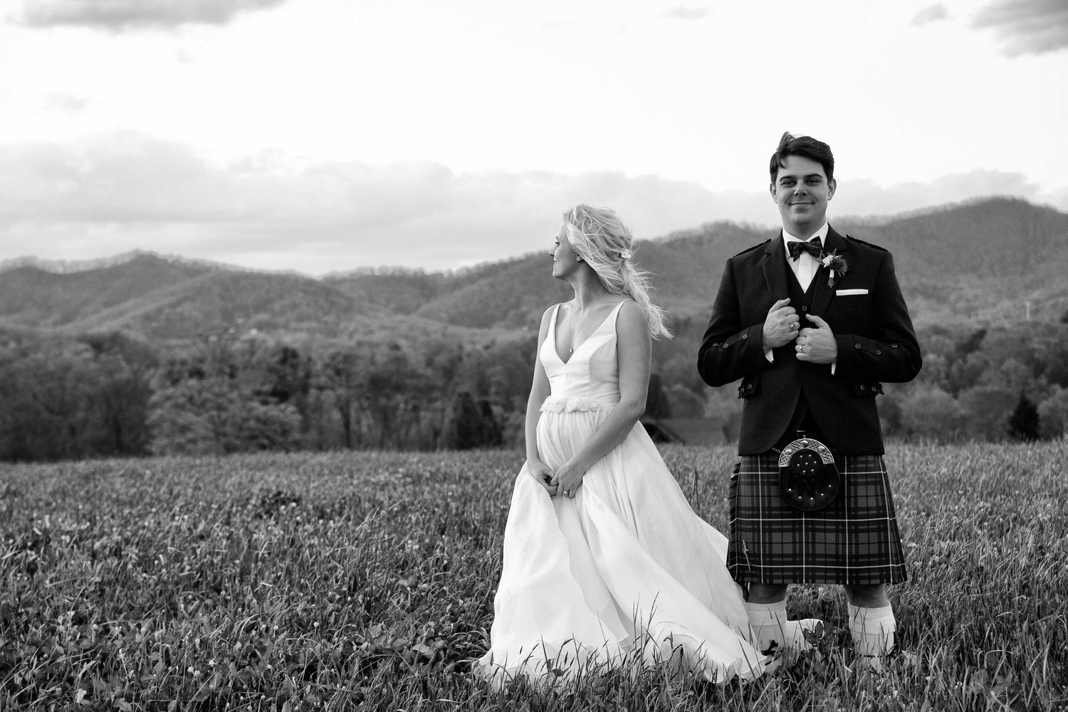 Black and white wedding photo of bride in gown and groom in Scottish kilt standing in field with mountain backdrop.