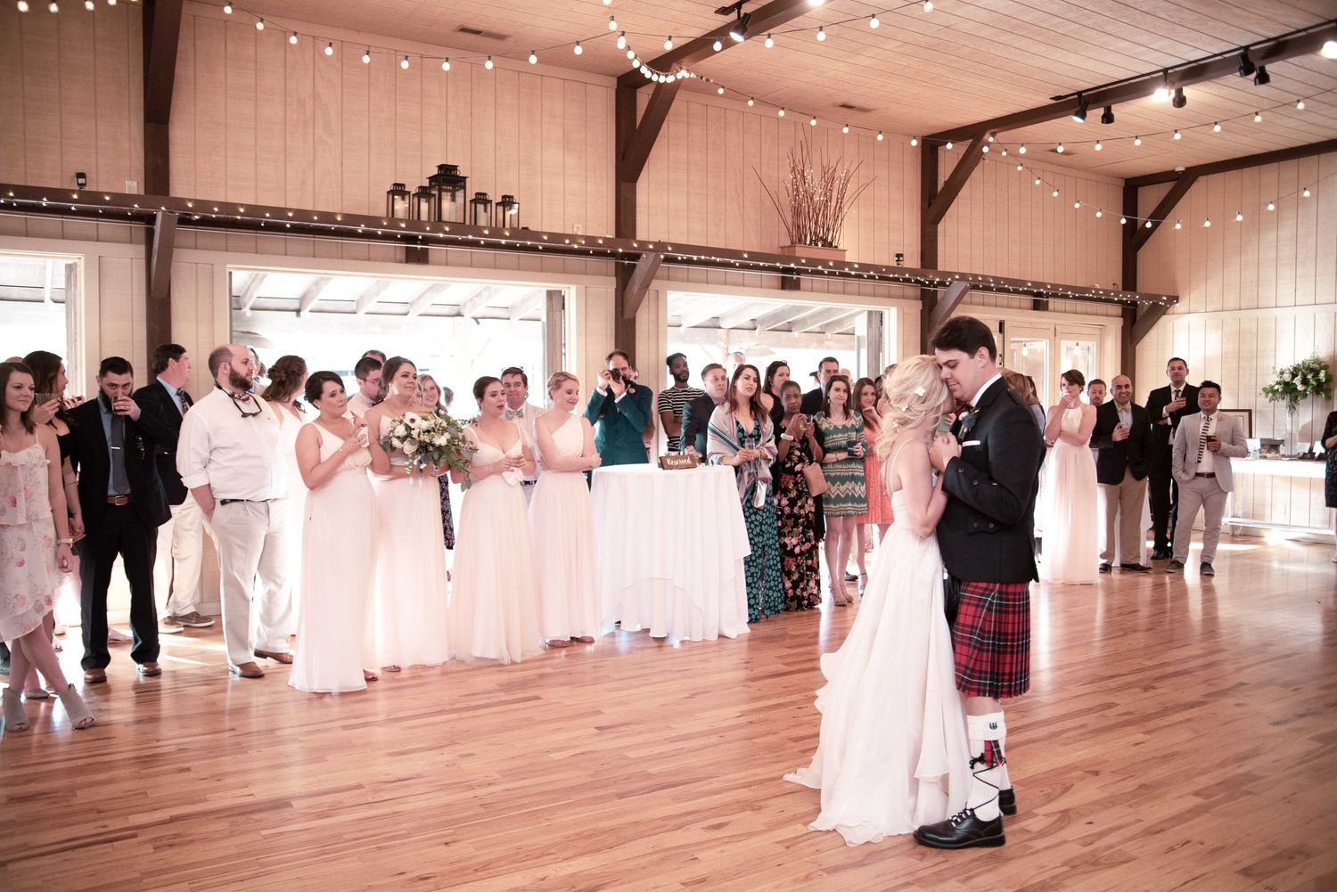 Bride and groom share first dance in rustic barn wedding venue with bridesmaids in white dresses and string lights.