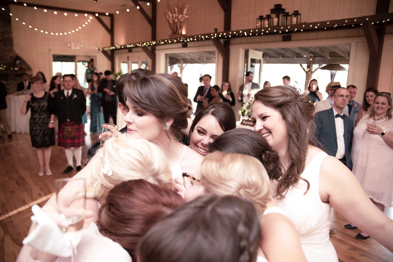 Bridesmaids in white dresses share a joyful group hug on the dance floor at a rustic barn wedding reception.