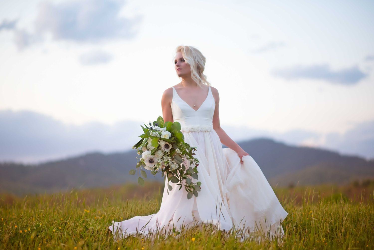 Blonde bride in white gown holds lush bouquet in mountain meadow at dusk.