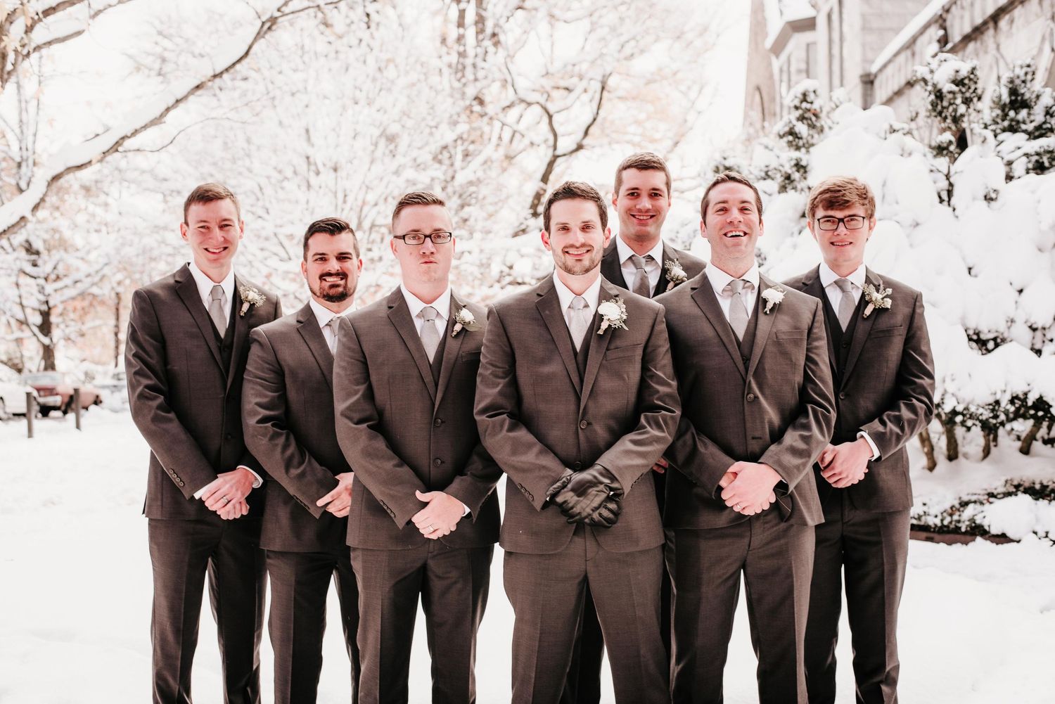 A group of seven groomsmen in matching brown suits posing outdoors in a snowy winter wedding setting.