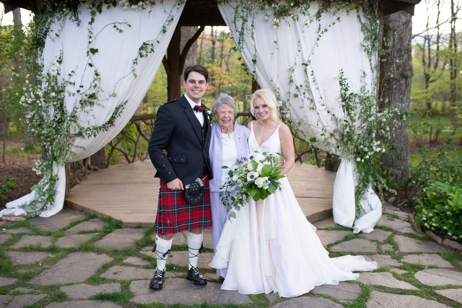 Bride and groom in Scottish kilt pose with officiant under floral arch at outdoor wedding ceremony.