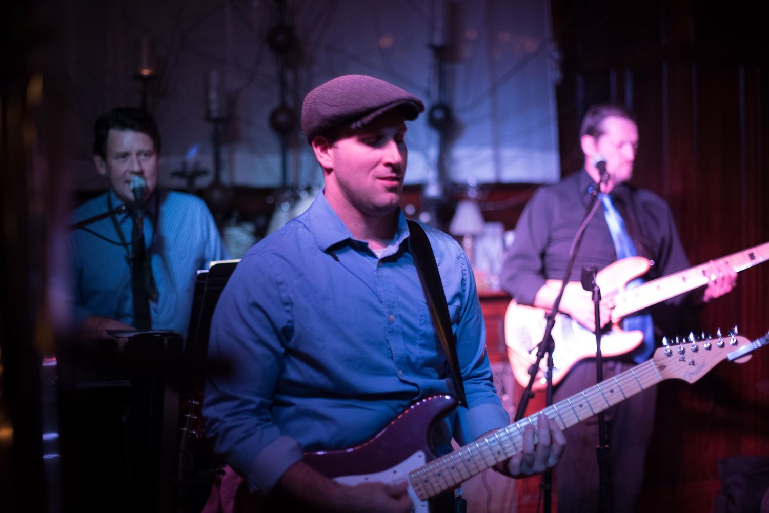 Three musicians performing live, with a guitarist in foreground wearing a cap, playing electric guitar on a dimly lit stage.