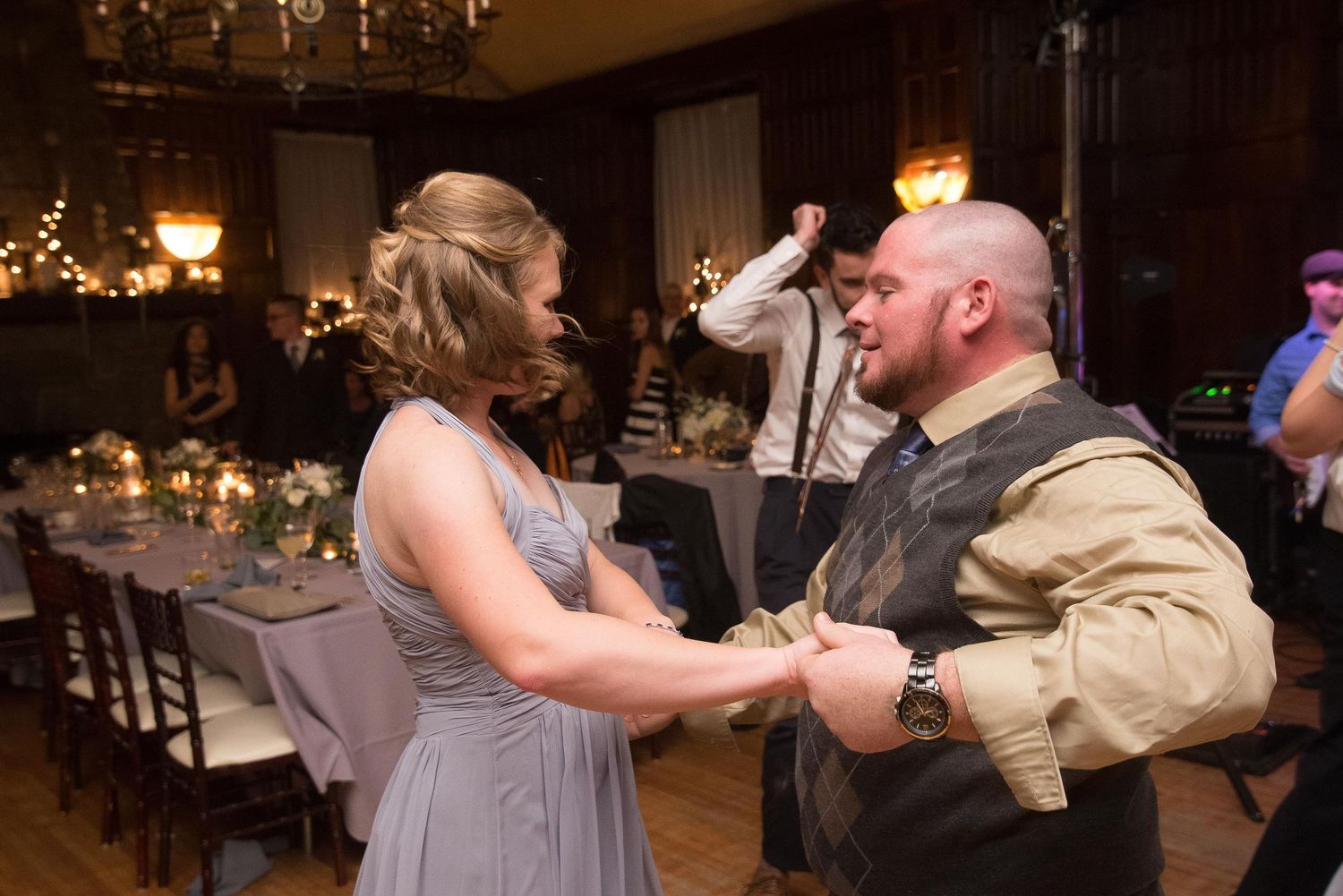 Couple dancing at a wedding reception in an elegant ballroom with candlelit tables and guests in the background.