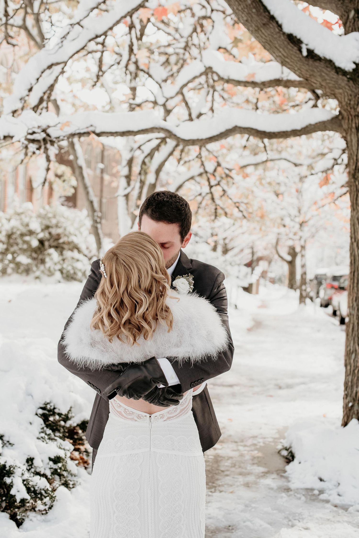 Bride and groom embrace in a snowy winter wonderland, with snow-covered trees lining the street behind them.
