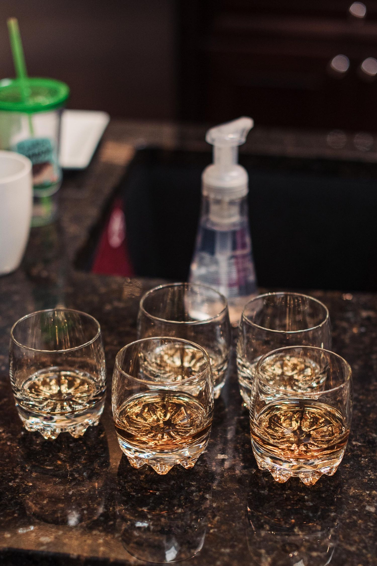 Five crystal whiskey glasses with amber liquid on a dark granite countertop near a kitchen sink.