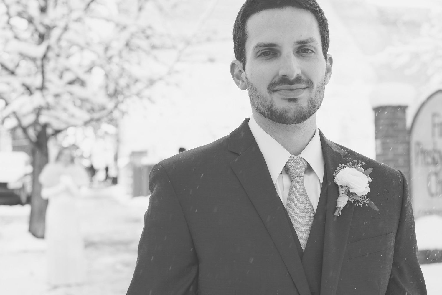 Groom in dark suit with boutonniere poses outdoors in snowy winter setting on wedding day.