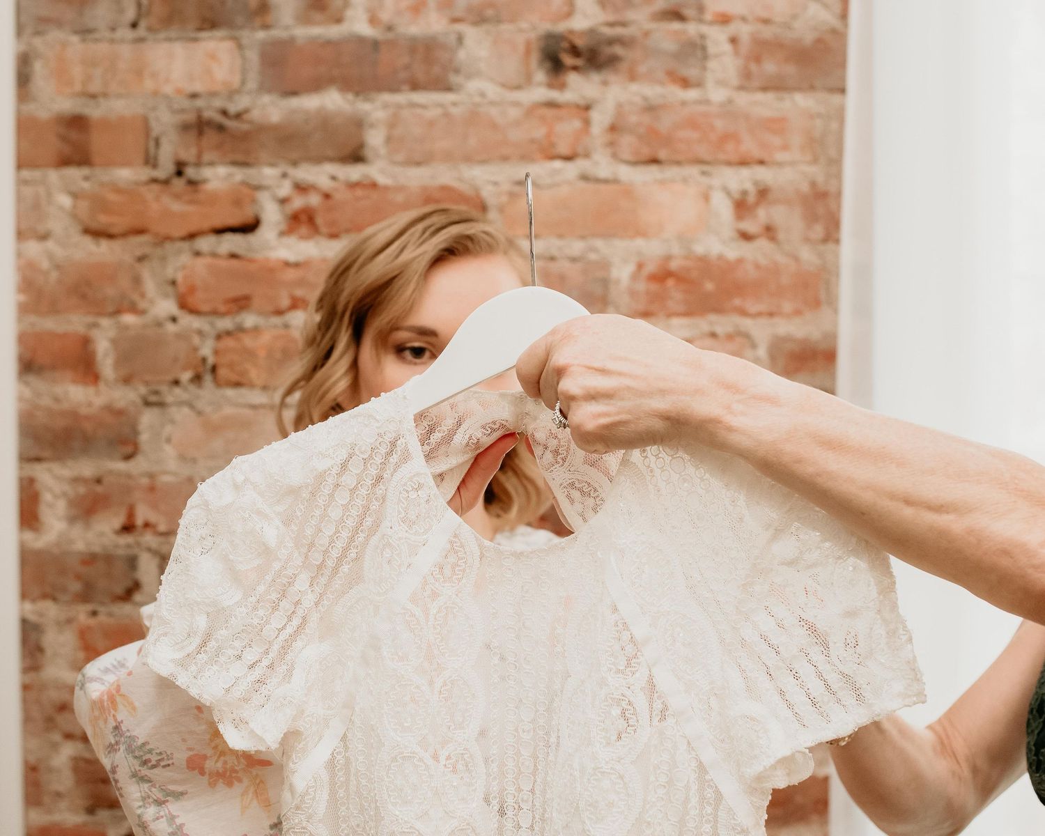 Woman holding a white lace wedding dress on a hanger against a rustic brick wall backdrop.