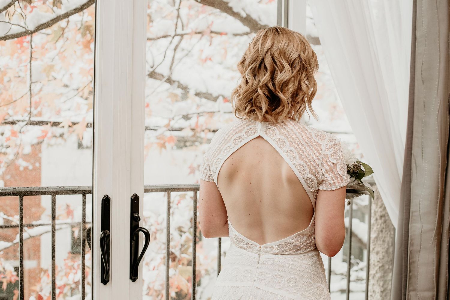 Bride in elegant backless lace wedding gown standing by French doors with autumn foliage backdrop.