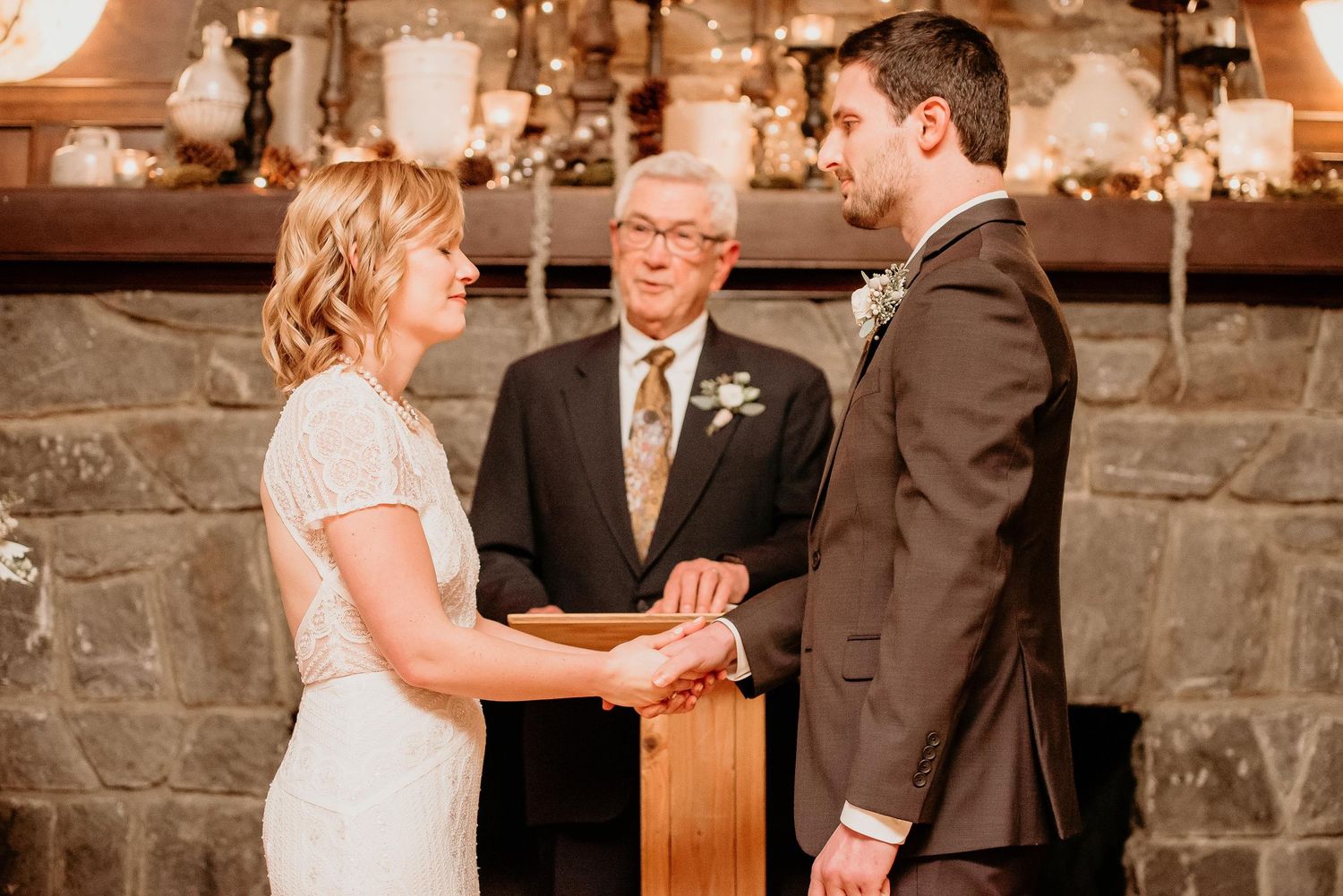 Bride and groom exchange vows holding hands before officiant at rustic stone fireplace wedding ceremony.