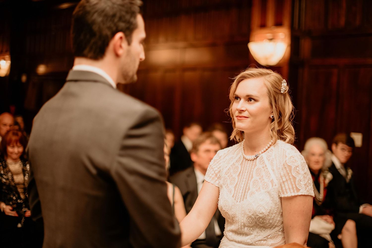 Bride smiling lovingly at groom during indoor wedding ceremony with guests in background.