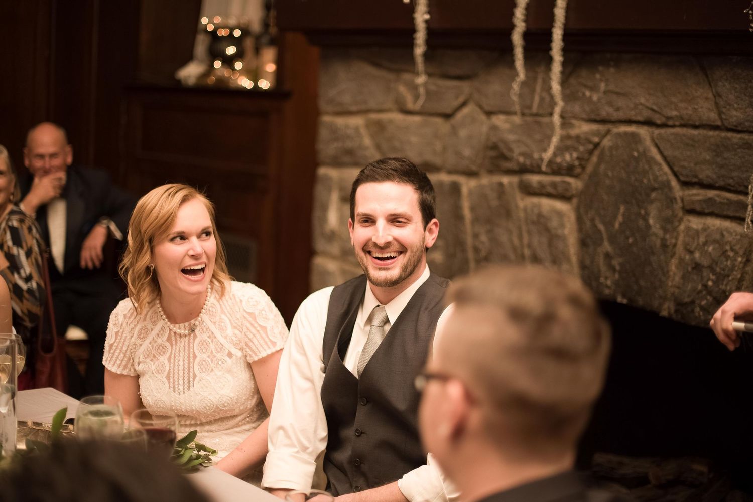 A smiling couple sits together at a wedding reception near a stone fireplace in a warmly lit venue.