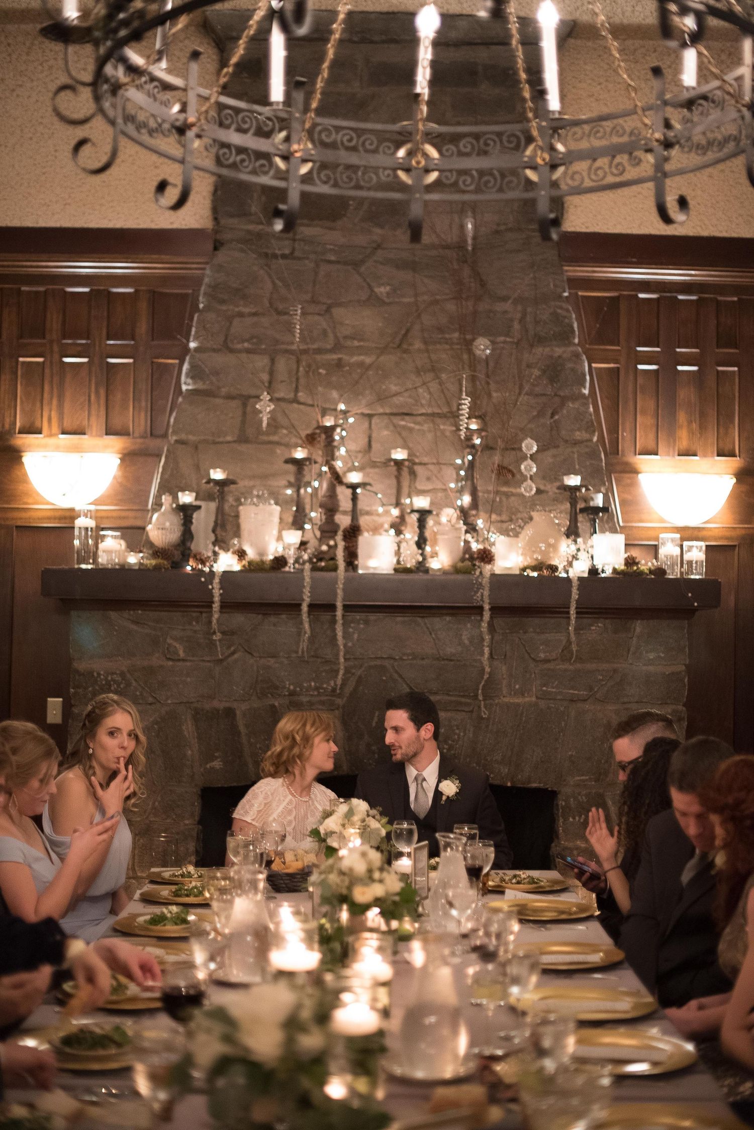 Couple shares intimate wedding dinner by candlelit stone fireplace, surrounded by guests at elegantly set tables.