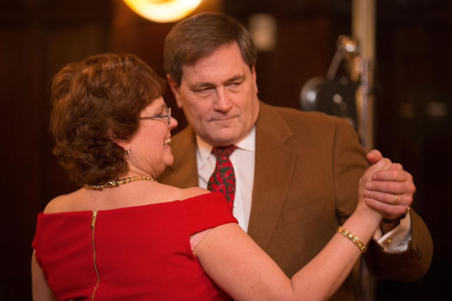 Elegant couple dancing together, woman in red off-shoulder dress, man in suit with red tie, at a formal event.