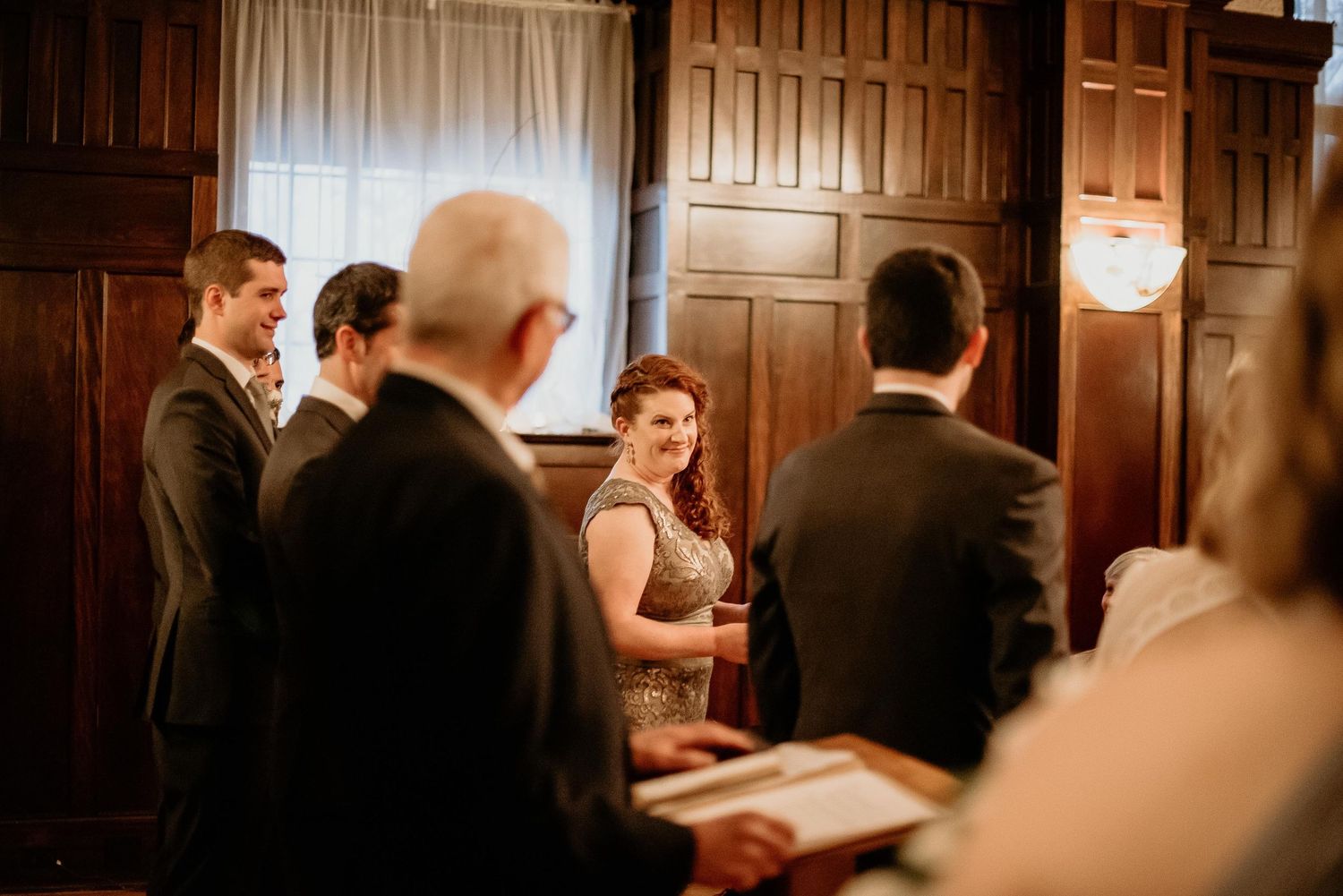 Bride smiling during an intimate wedding ceremony in an elegant wood-paneled room with guests gathered around.