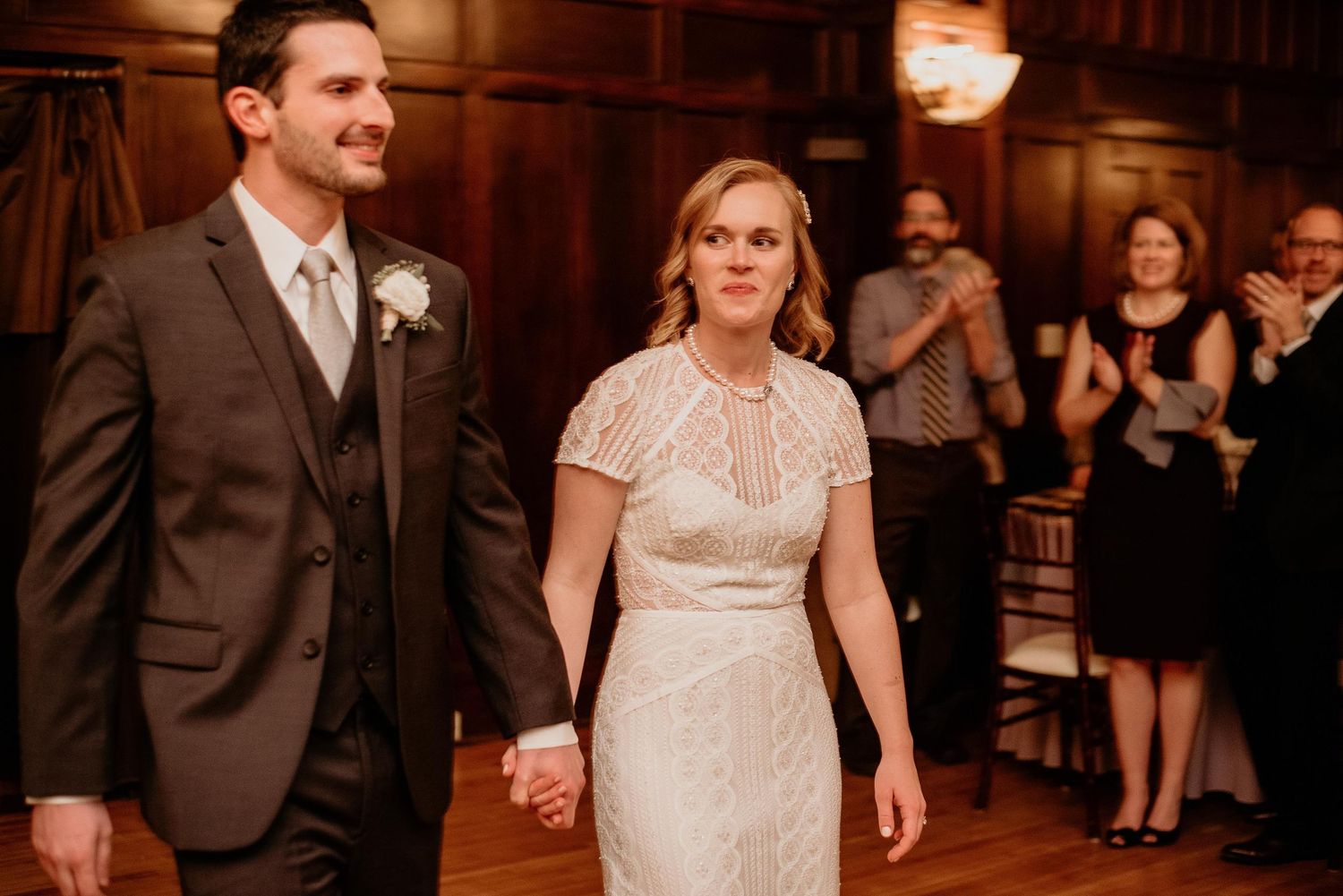 Bride and groom holding hands walking into wedding reception in elegant wood-paneled venue with guests applauding behind them.