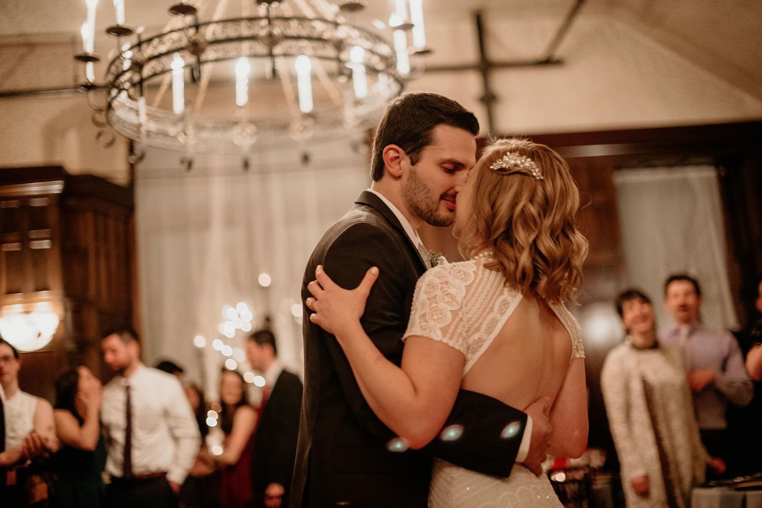 Bride and groom share a romantic first dance kiss under a chandelier at their elegant wedding reception.