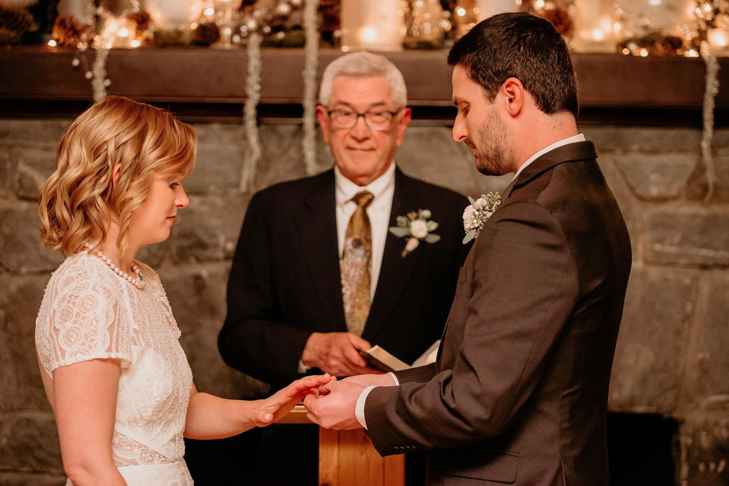 Bride and groom exchange rings before officiant at romantic indoor wedding ceremony by stone fireplace.