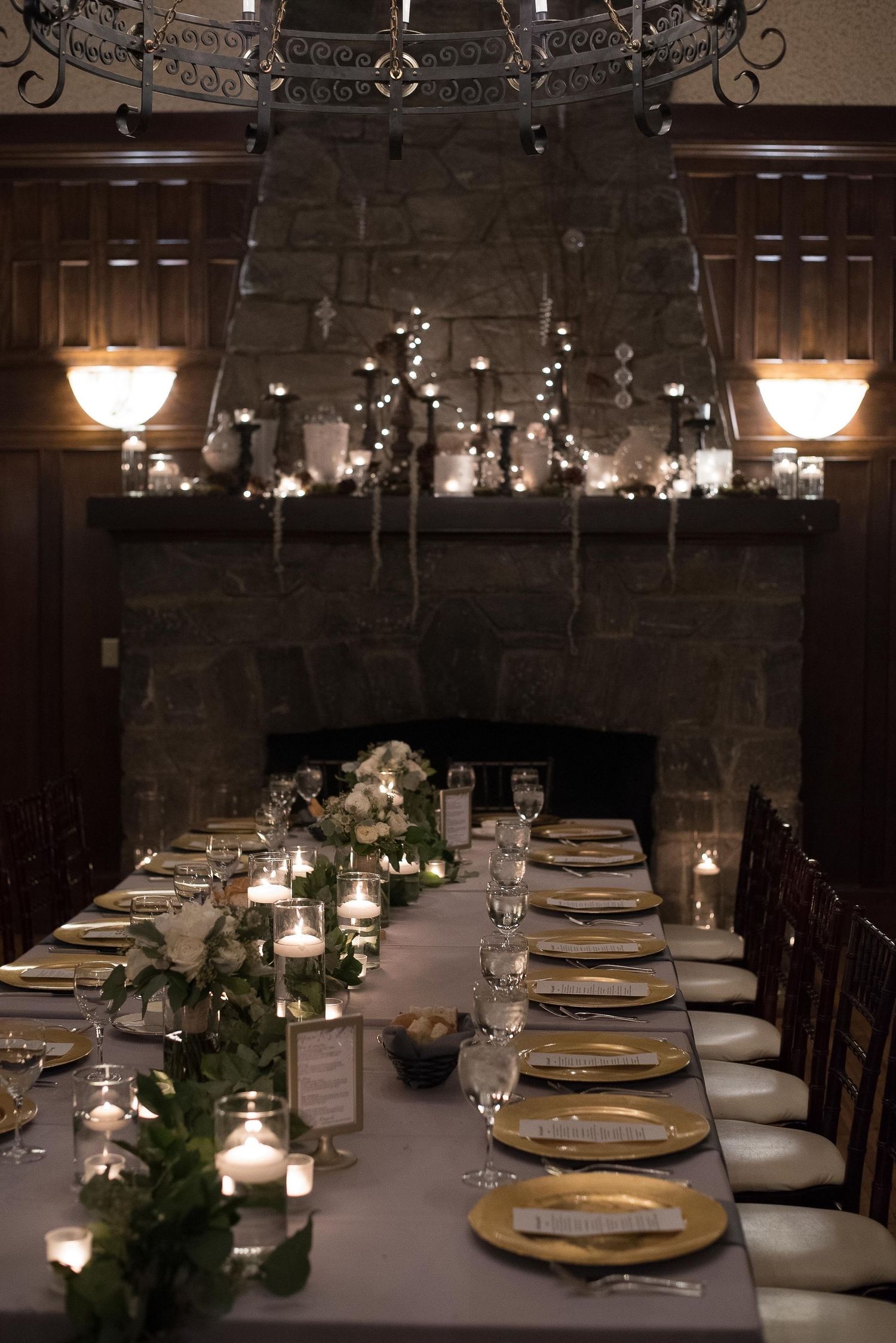 Elegant candlelit dining table with gold chargers, greenery runner, and stone fireplace in a dark, romantic wedding reception setting.