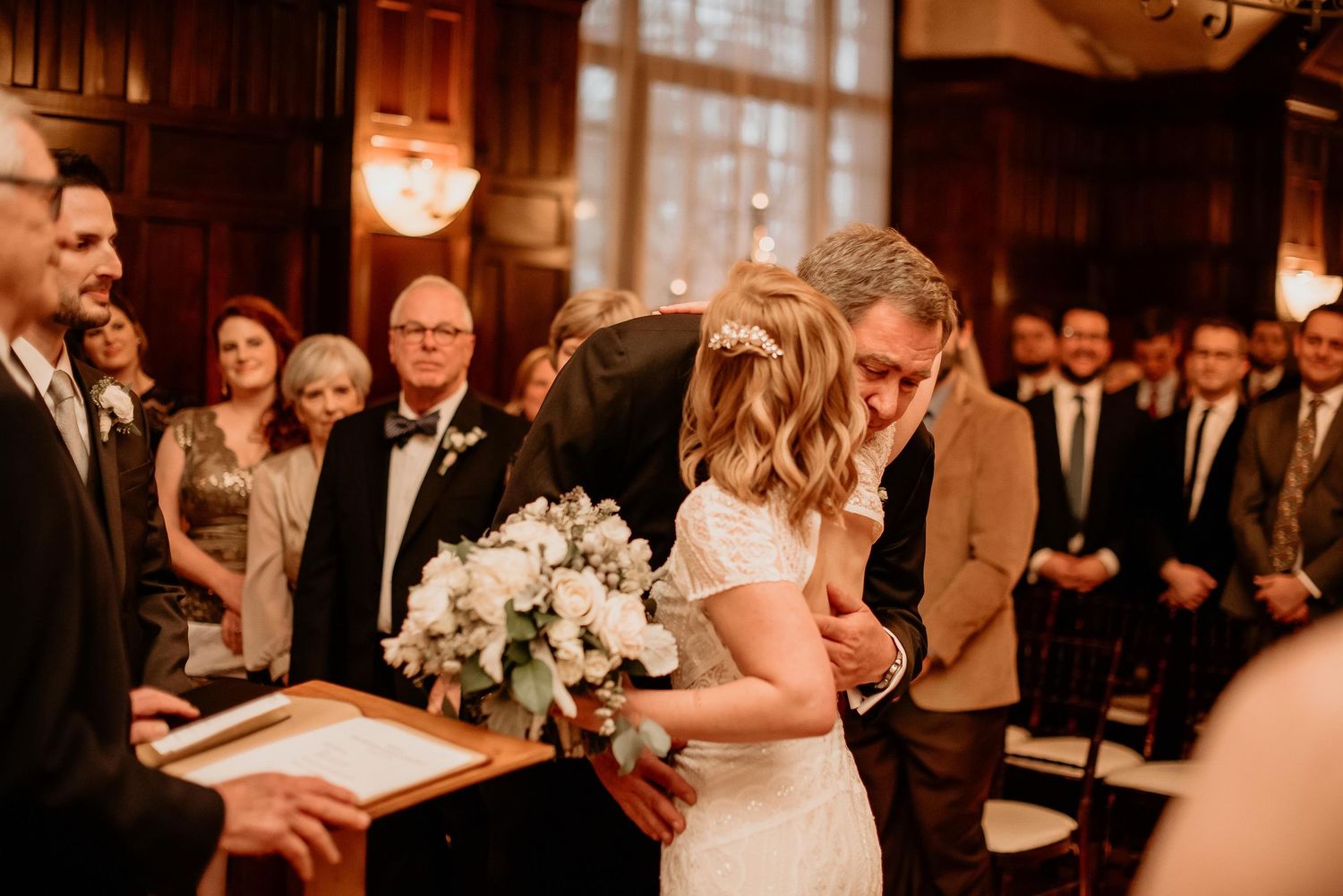 Bride and groom share emotional first kiss at indoor wedding ceremony with guests watching in elegant wood-paneled venue.