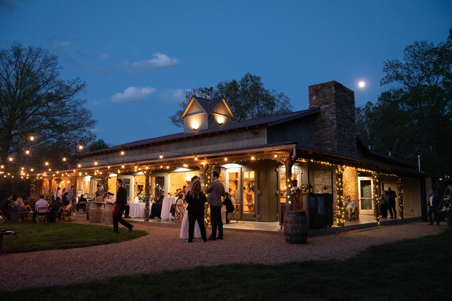 Rustic wedding venue glowing with string lights at dusk, guests mingling outside under a moonlit sky.