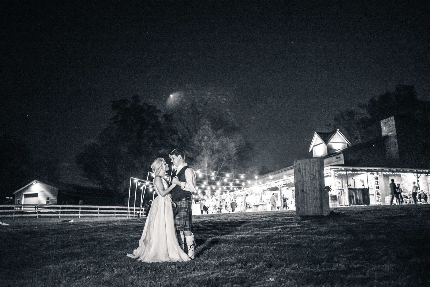 Black and white wedding photo of couple embracing outdoors at night with illuminated barn venue in background.