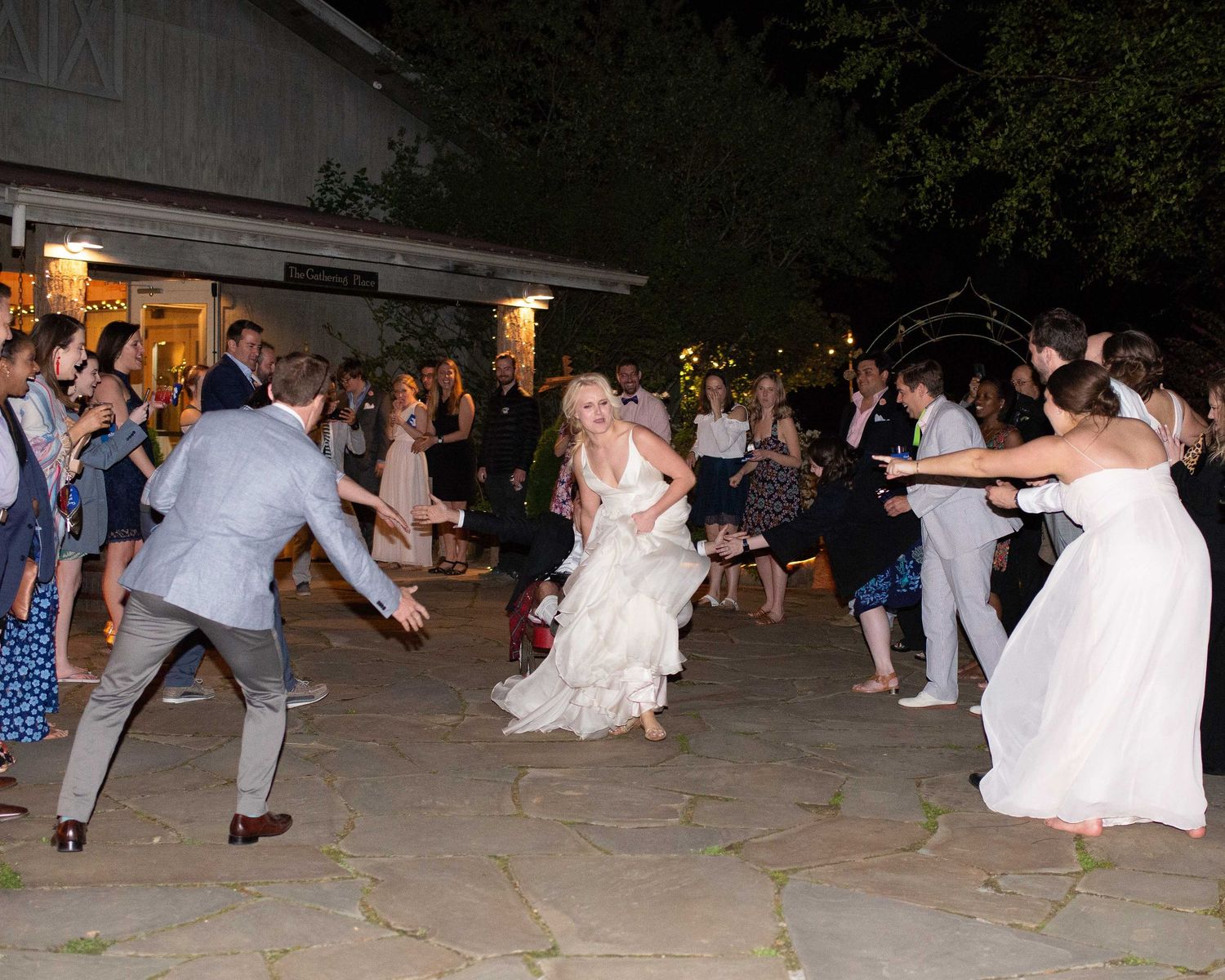 Bride and groom dancing joyfully with wedding guests on outdoor stone patio at night during reception celebration.