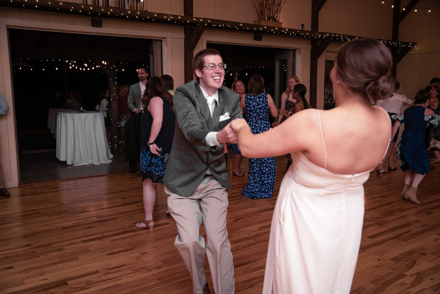 Couple dancing joyfully at a wedding reception on a wooden dance floor with string lights in the background.