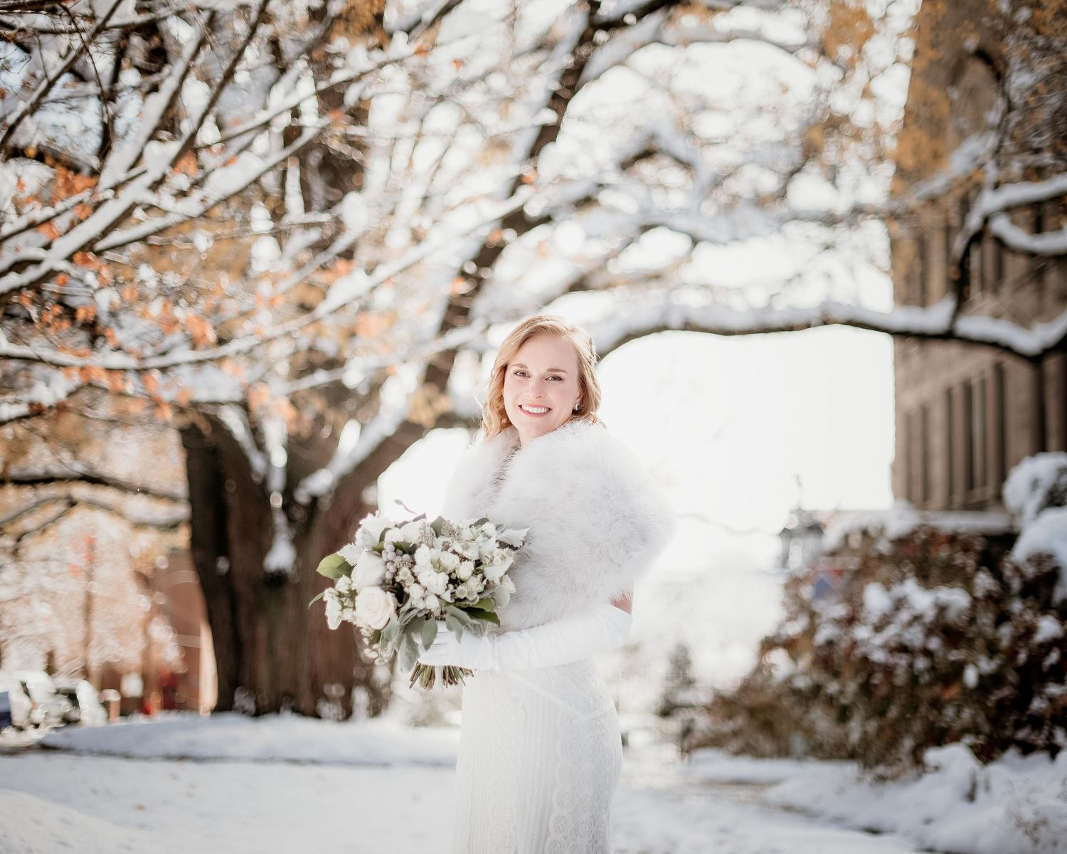 Smiling bride in white fur stole holding bouquet outdoors in snowy winter wonderland setting.