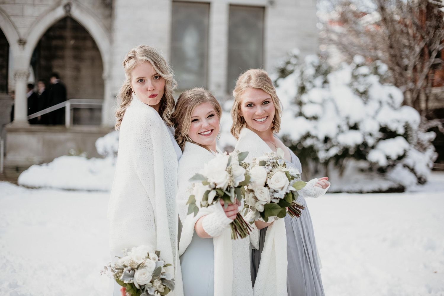 Three bridesmaids in white wraps holding bouquets pose outdoors in a snowy winter wedding setting.