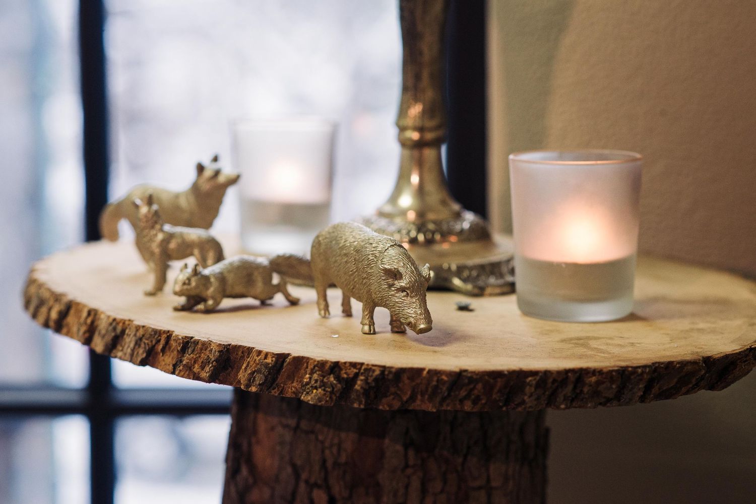 Gold animal figurines and frosted candle holders arranged on a rustic wood slice table near a window.
