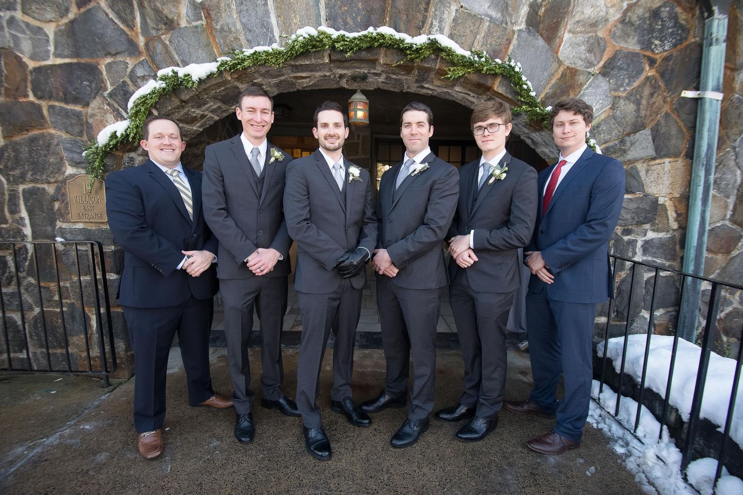 Six groomsmen in gray suits pose together outside a stone building decorated with greenery at a winter wedding.