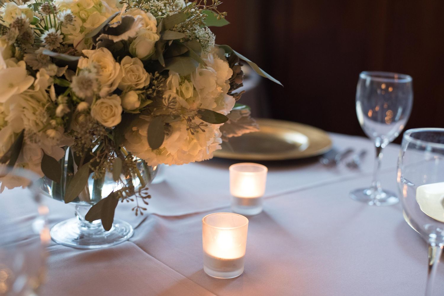 Elegant wedding table setting with white floral centerpiece, glowing candles, gold charger plates, and crystal wine glasses.