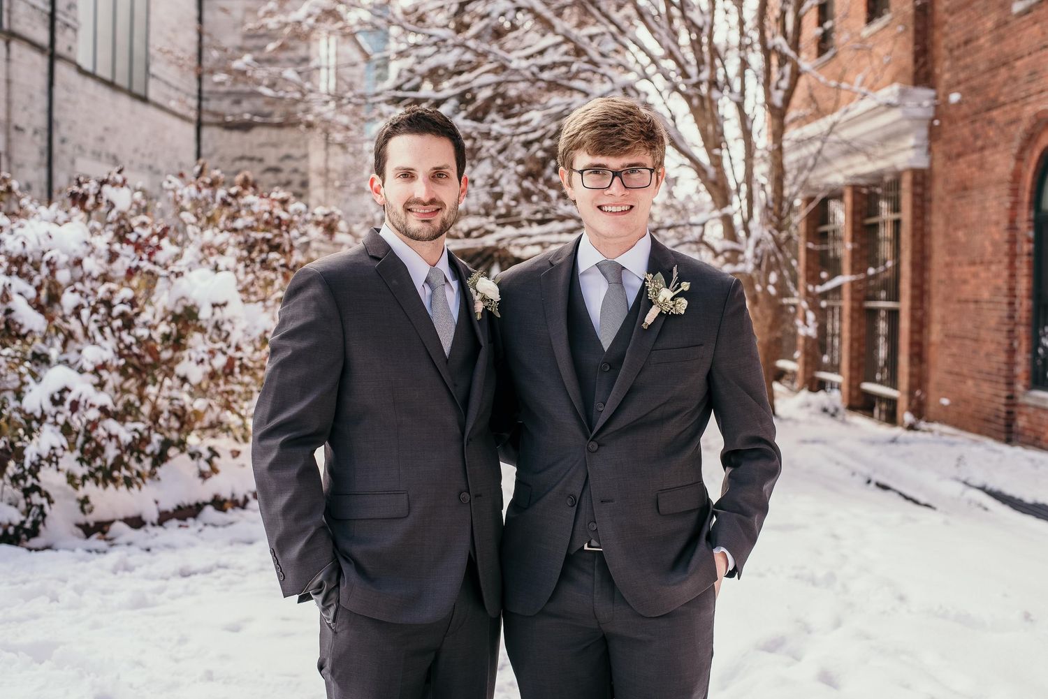 Two men in dark suits with boutonnieres pose outdoors in a snowy winter setting near a brick building.