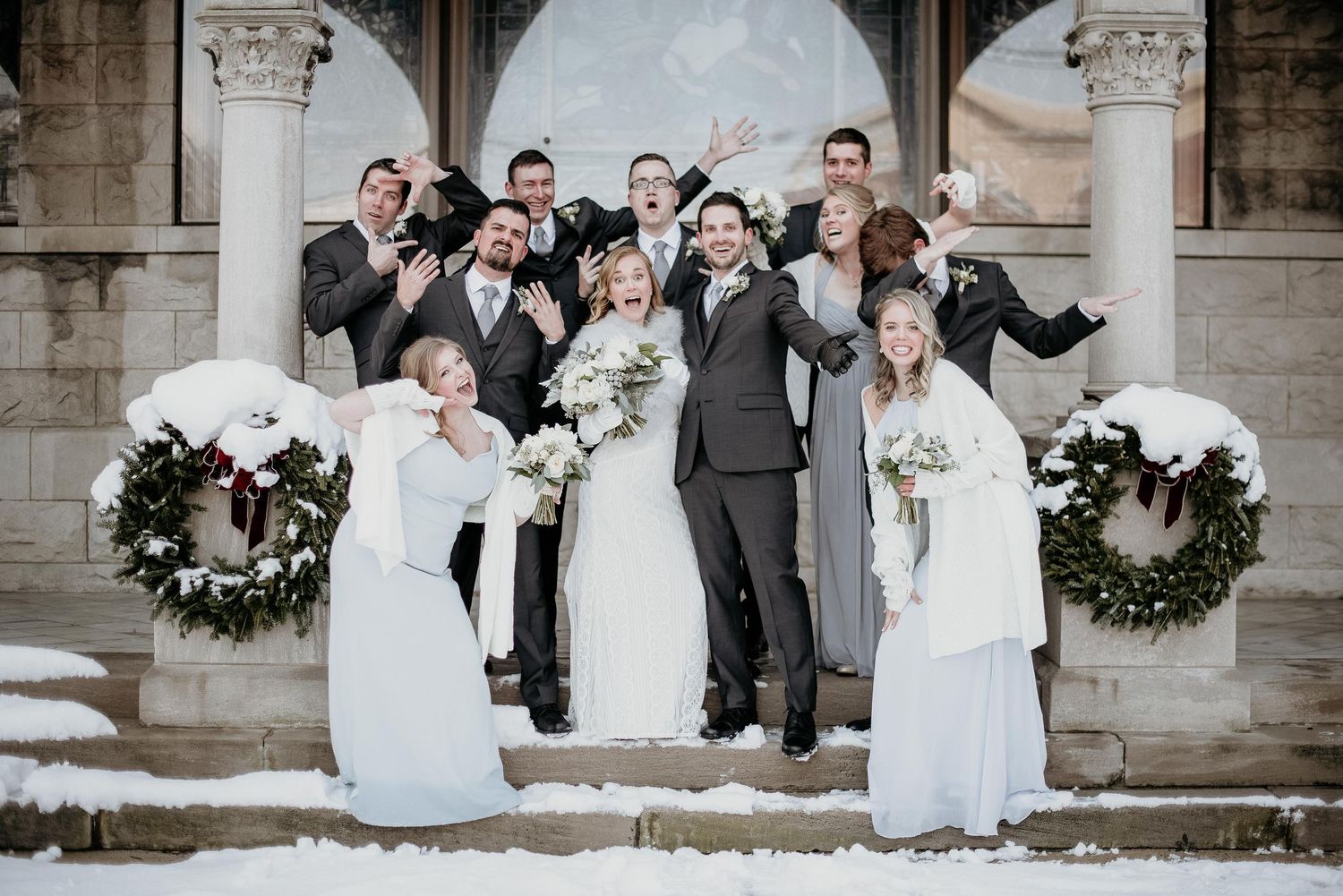 Wedding party celebrating on snowy steps with wreaths, bridesmaids in white gowns and groomsmen in gray suits jumping joyfully.