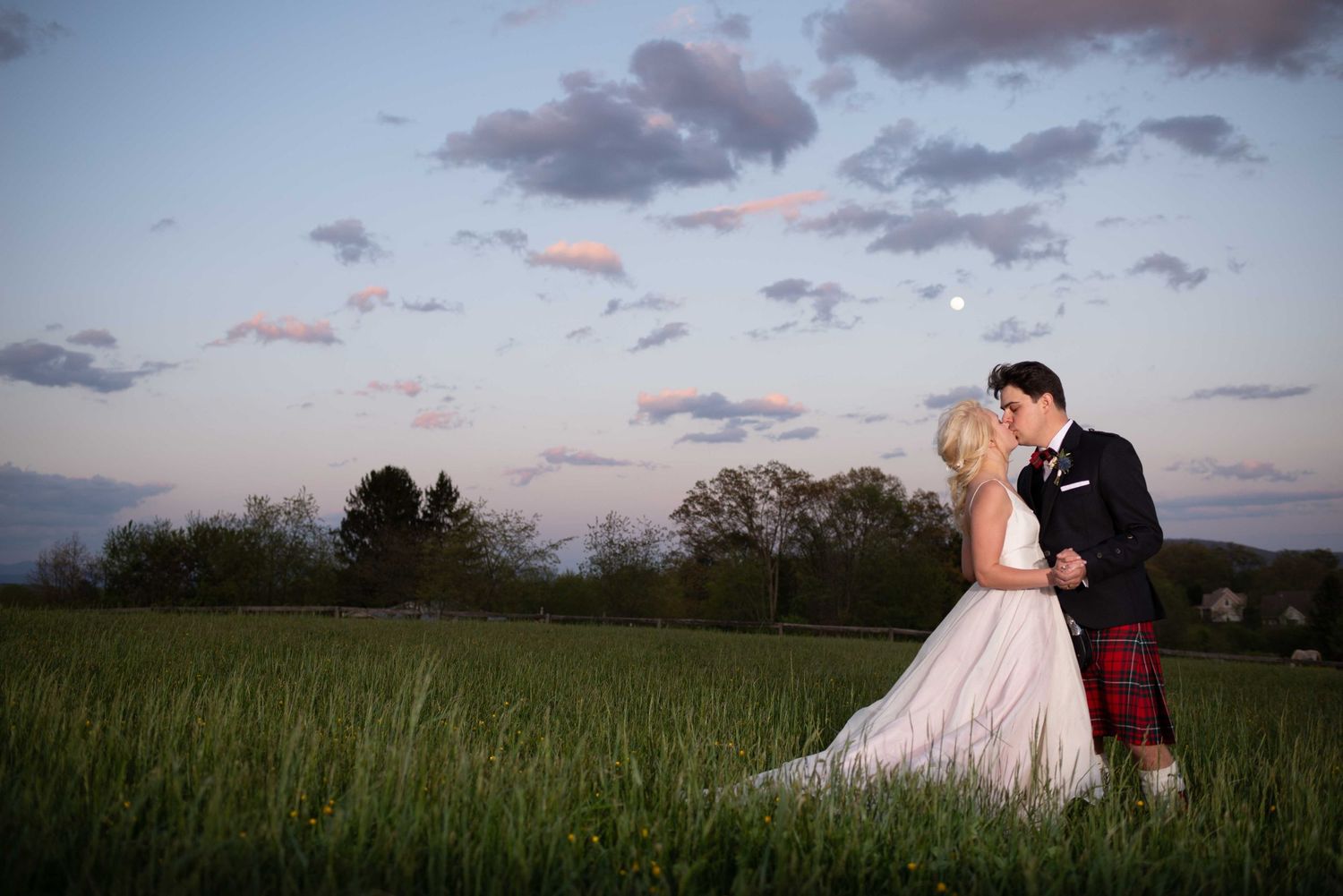 Bride and groom share a romantic kiss in a lush green field at sunset, groom wearing a red tartan kilt.