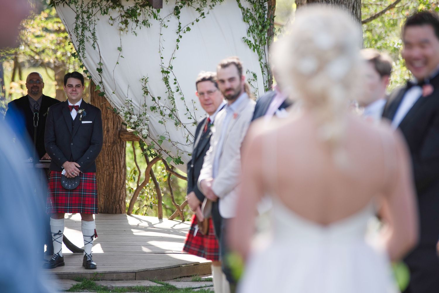 Groom in Scottish kilt watches bride walk down the aisle at an outdoor wedding ceremony.