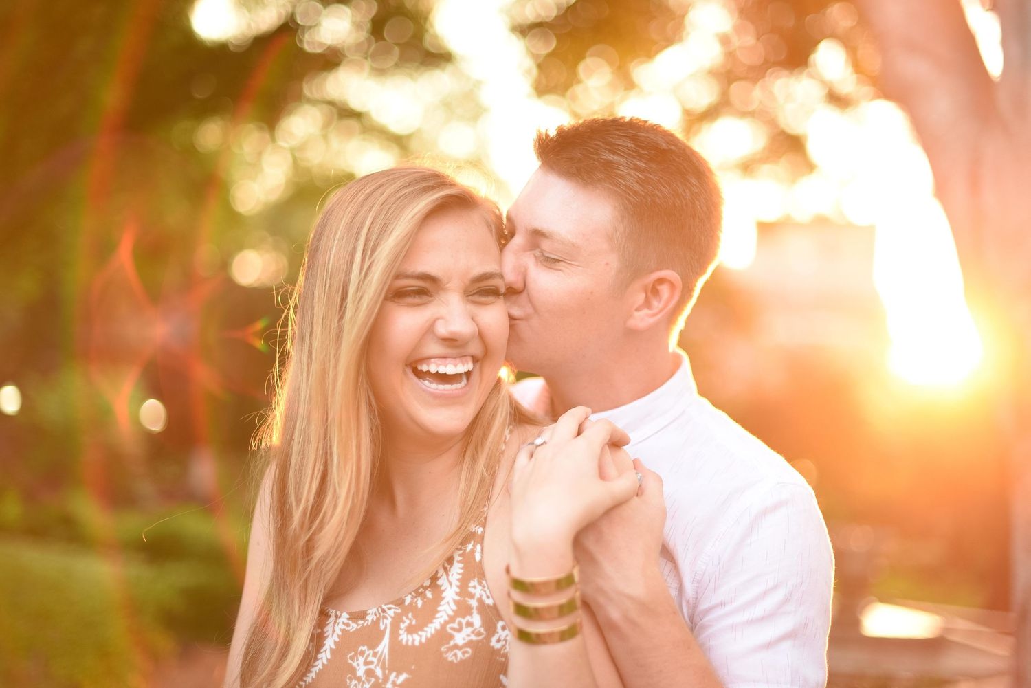 Joyful woman laughs as man kisses her cheek during a golden sunset engagement photo session outdoors.