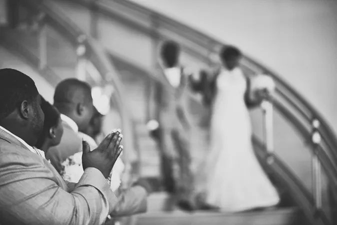 Black and white photo of wedding guests praying on stairs as bride and groom ascend in background.