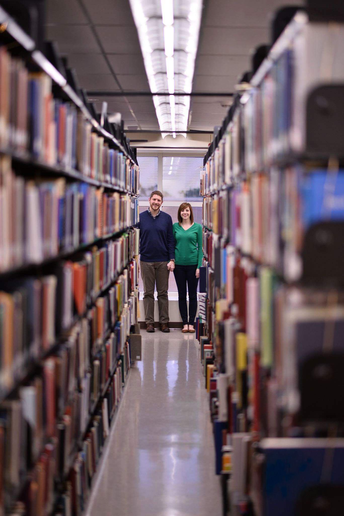 Happy couple holding hands while standing between tall bookshelves in a university library.