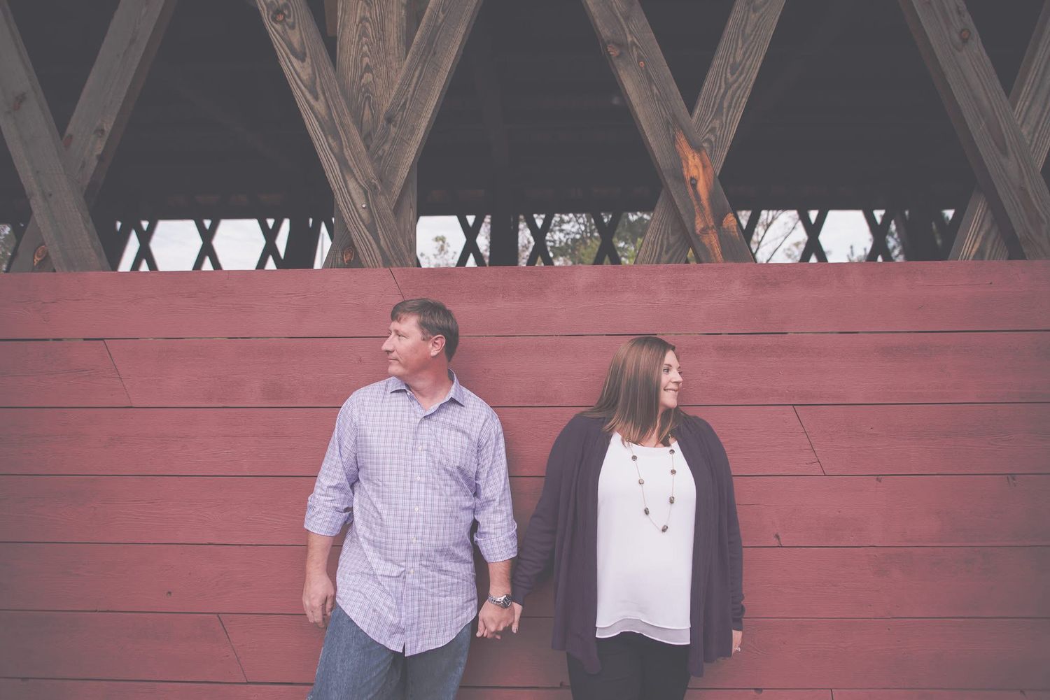 Couple holds hands while looking away from each other against a red wooden wall outdoors.