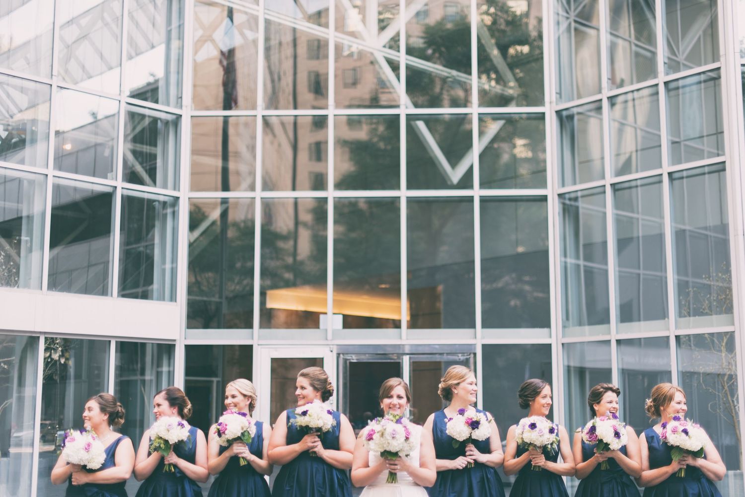 Bridesmaids in navy dresses hold bouquets while lined up before a large glass building facade.