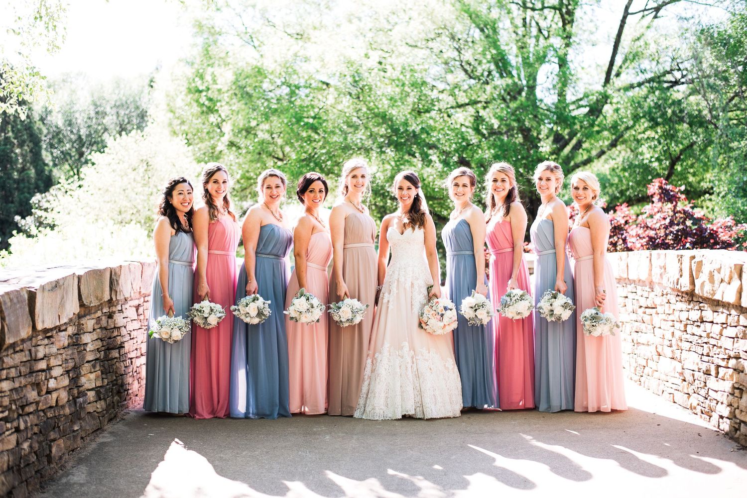 Bride poses with ten bridesmaids in mismatched pink and blue gowns holding white bouquets outdoors.