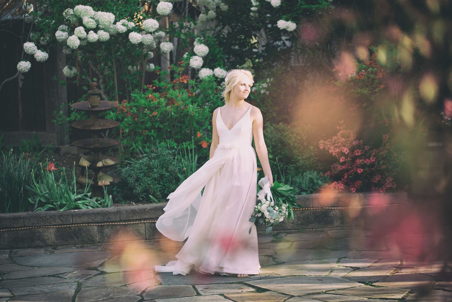 Bride in flowing pink gown walks through lush garden holding white bouquet.
