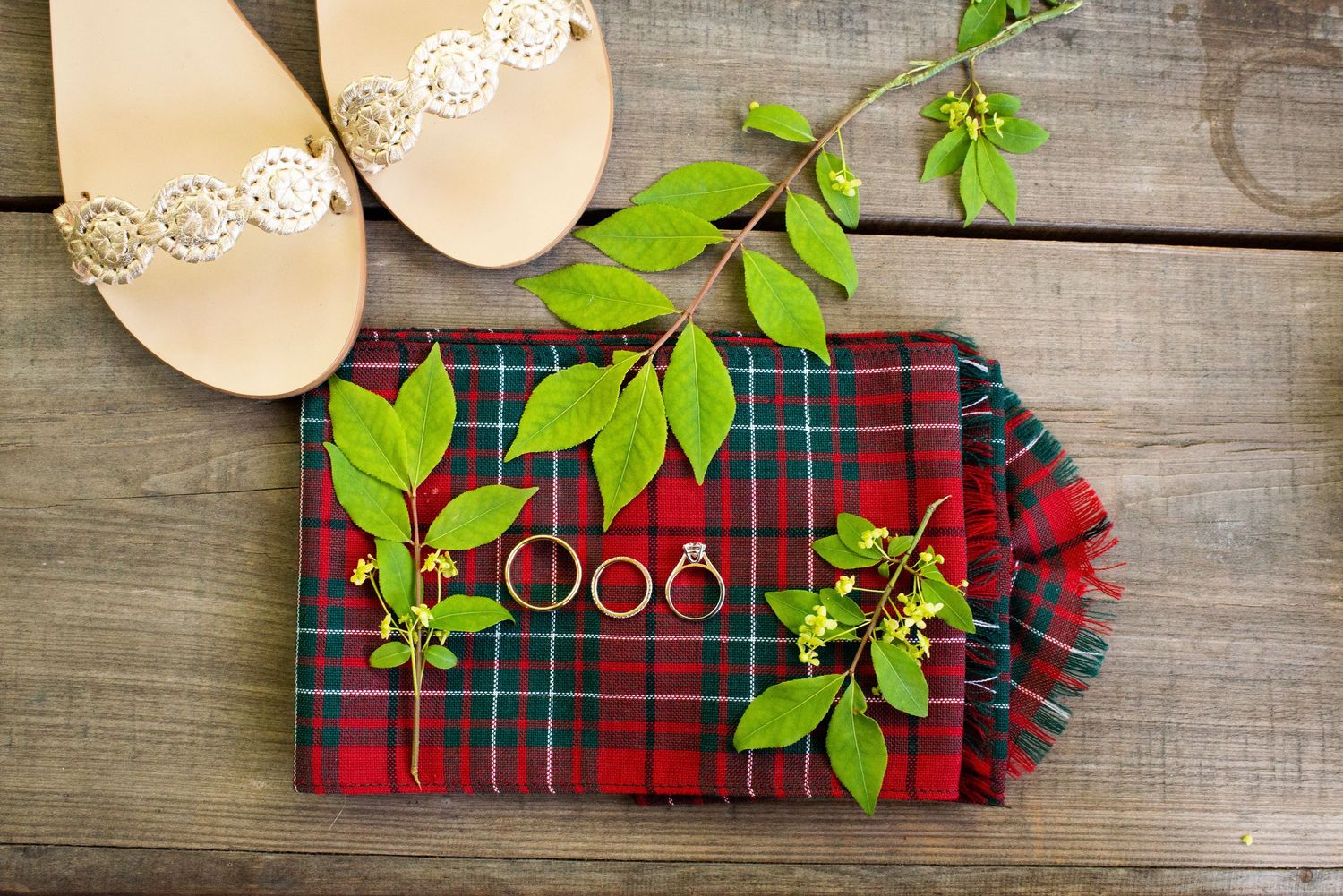 Wedding flat lay featuring gold sandals, rings, and green branches on a red tartan plaid fabric on a wooden surface.