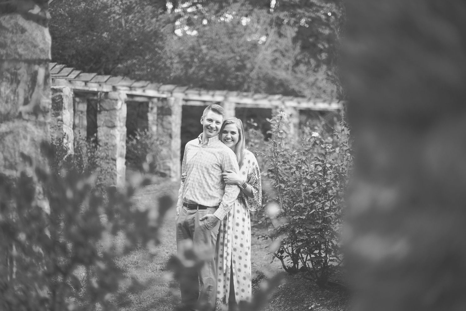 Black and white photo of a couple embracing in a lush garden near a stone wall.
