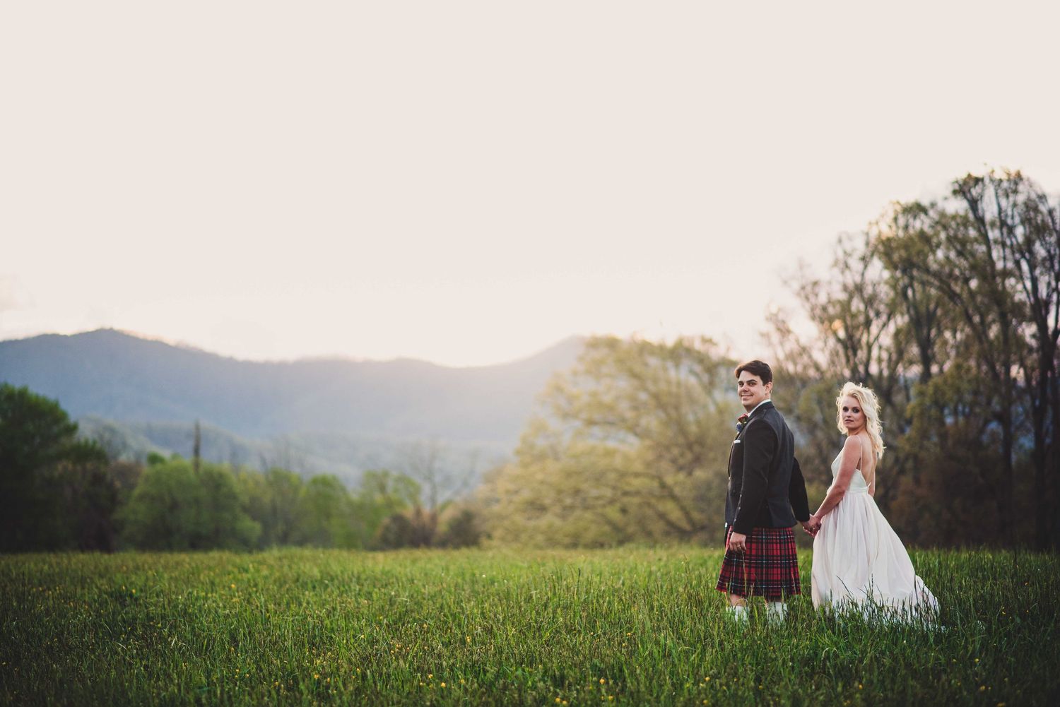 Bride and groom holding hands in a lush green field with mountains and trees in the background at sunset.