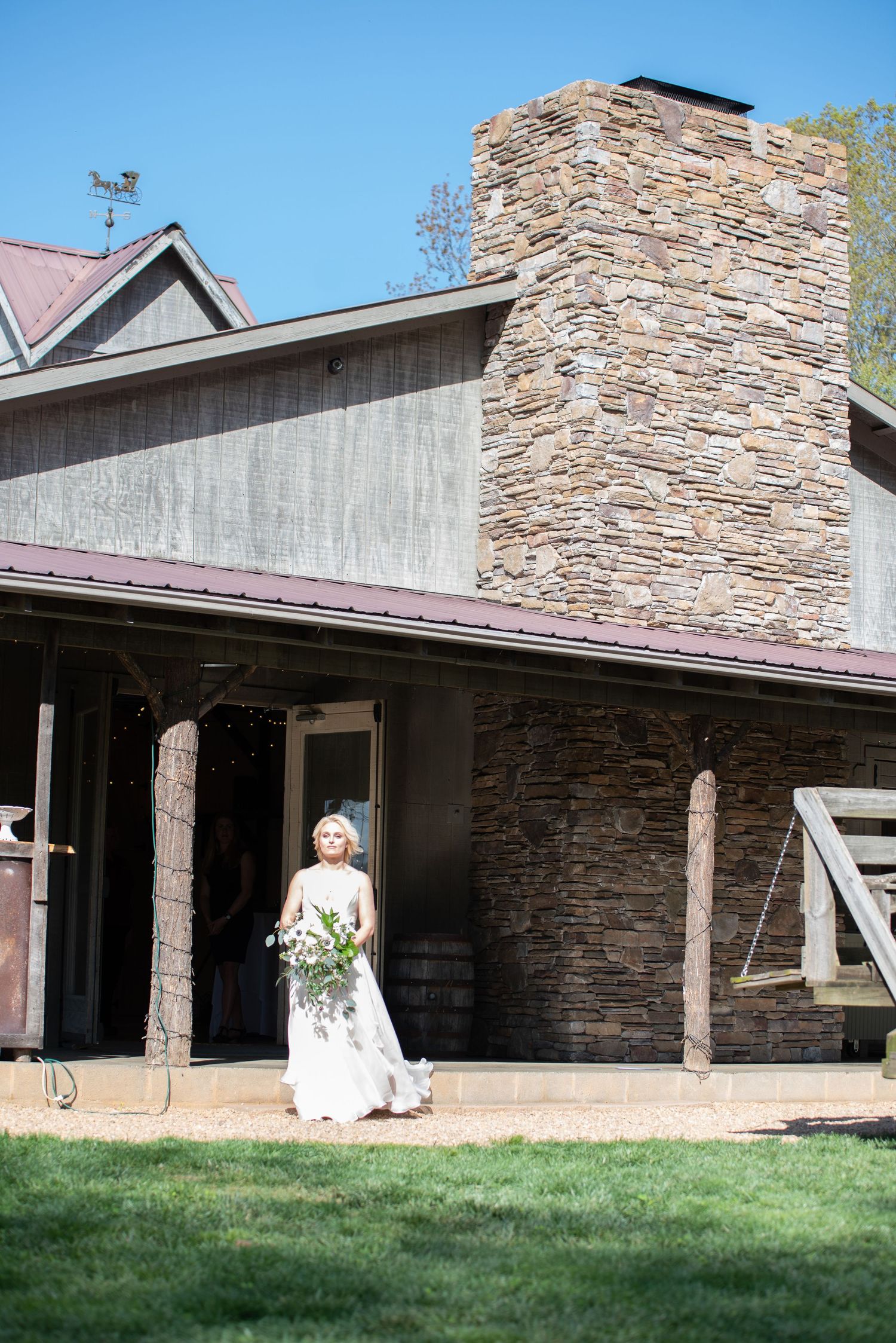 Blonde bride in elegant white gown holds bouquet outside rustic barn wedding venue.