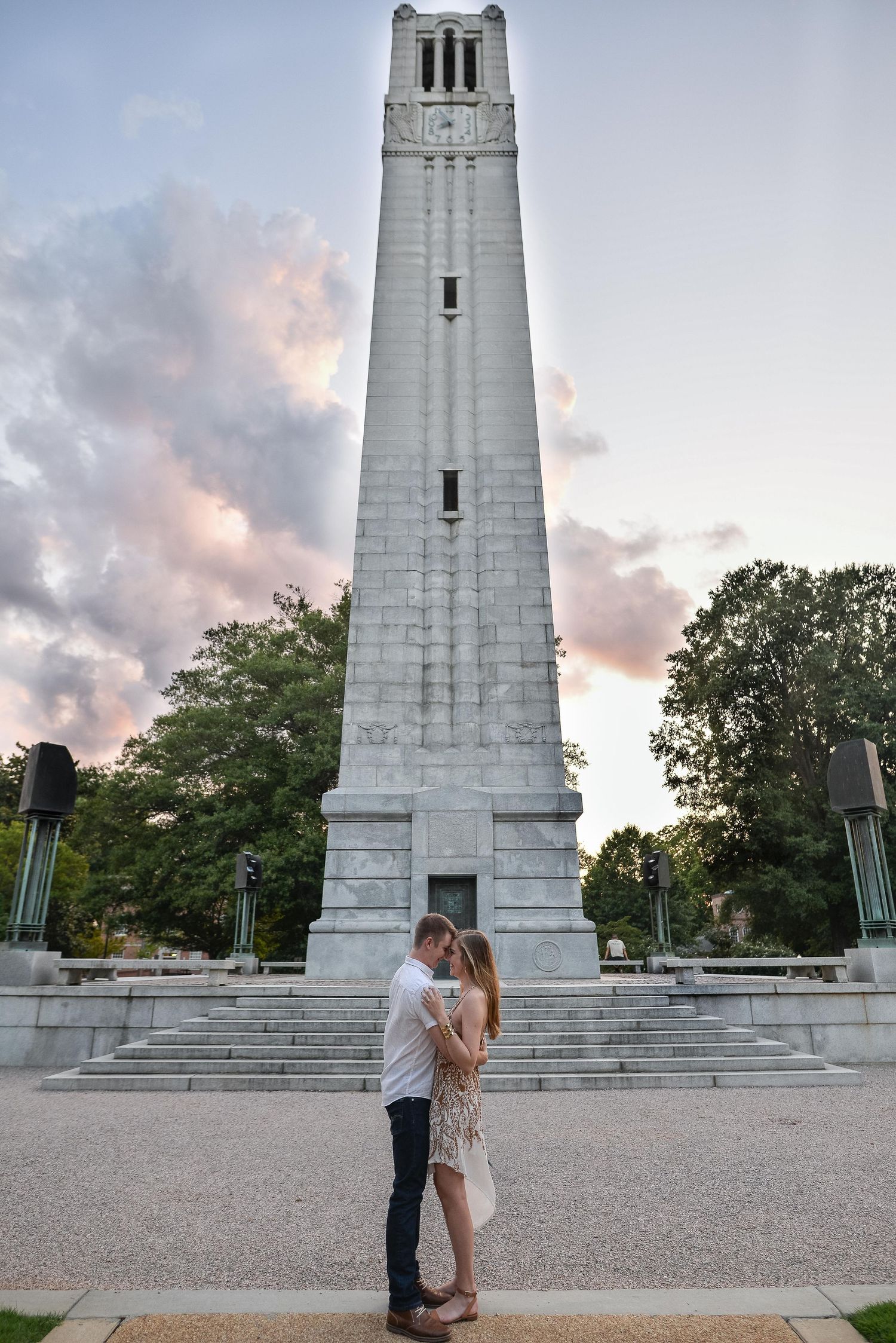 Couple shares a kiss in front of a tall stone bell tower at sunset with dramatic clouds above.