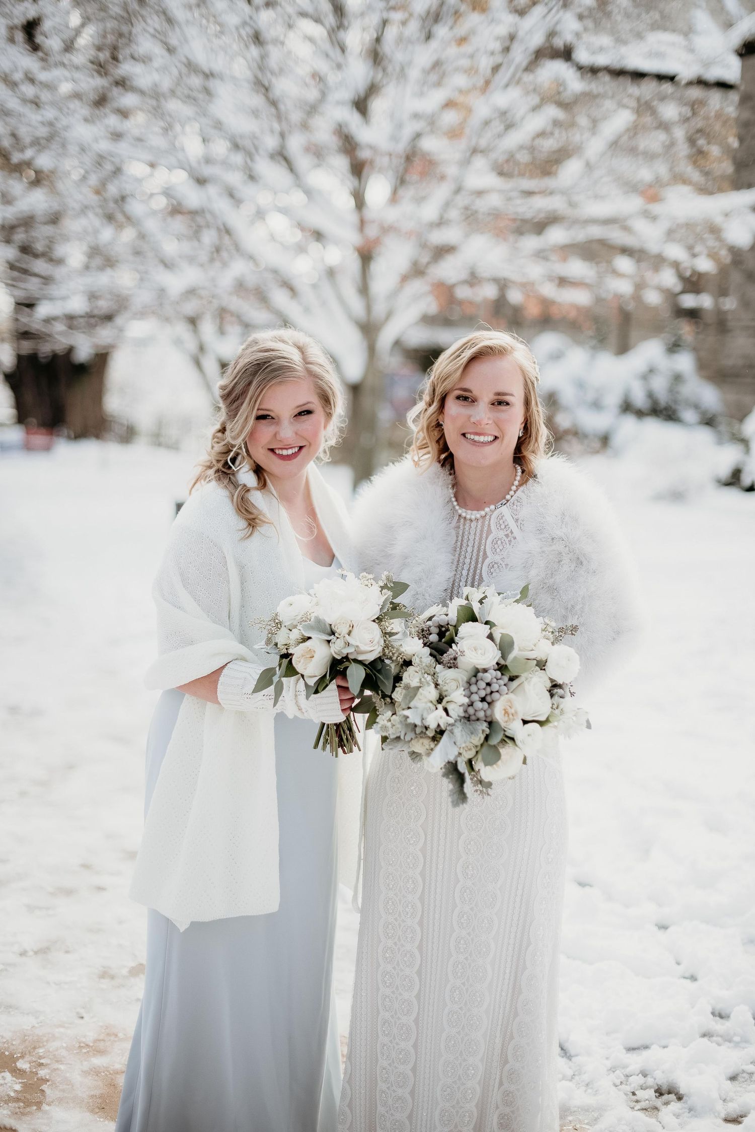 Two brides in white gowns and fur wraps hold bouquets outdoors in a snowy winter wedding setting.